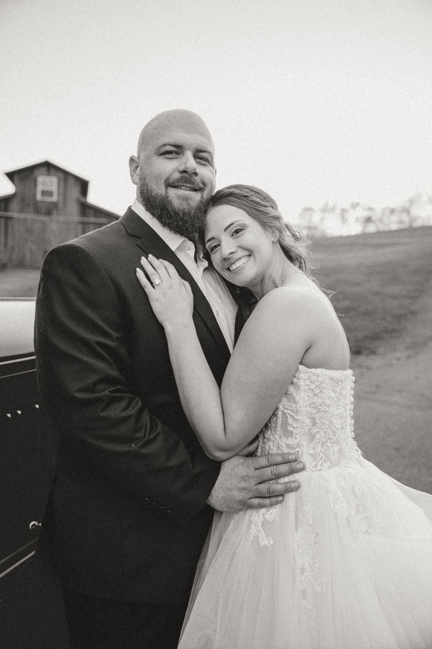 Black and white portrait of bearded groom and smiling bride embracing outdoors leaning against vintage vehicle.