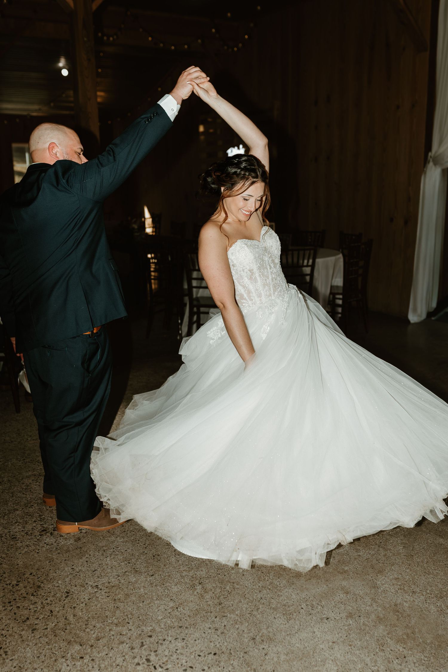Bride in white ballgown twirls during first dance with groom in dark suit at rustic barn wedding reception.