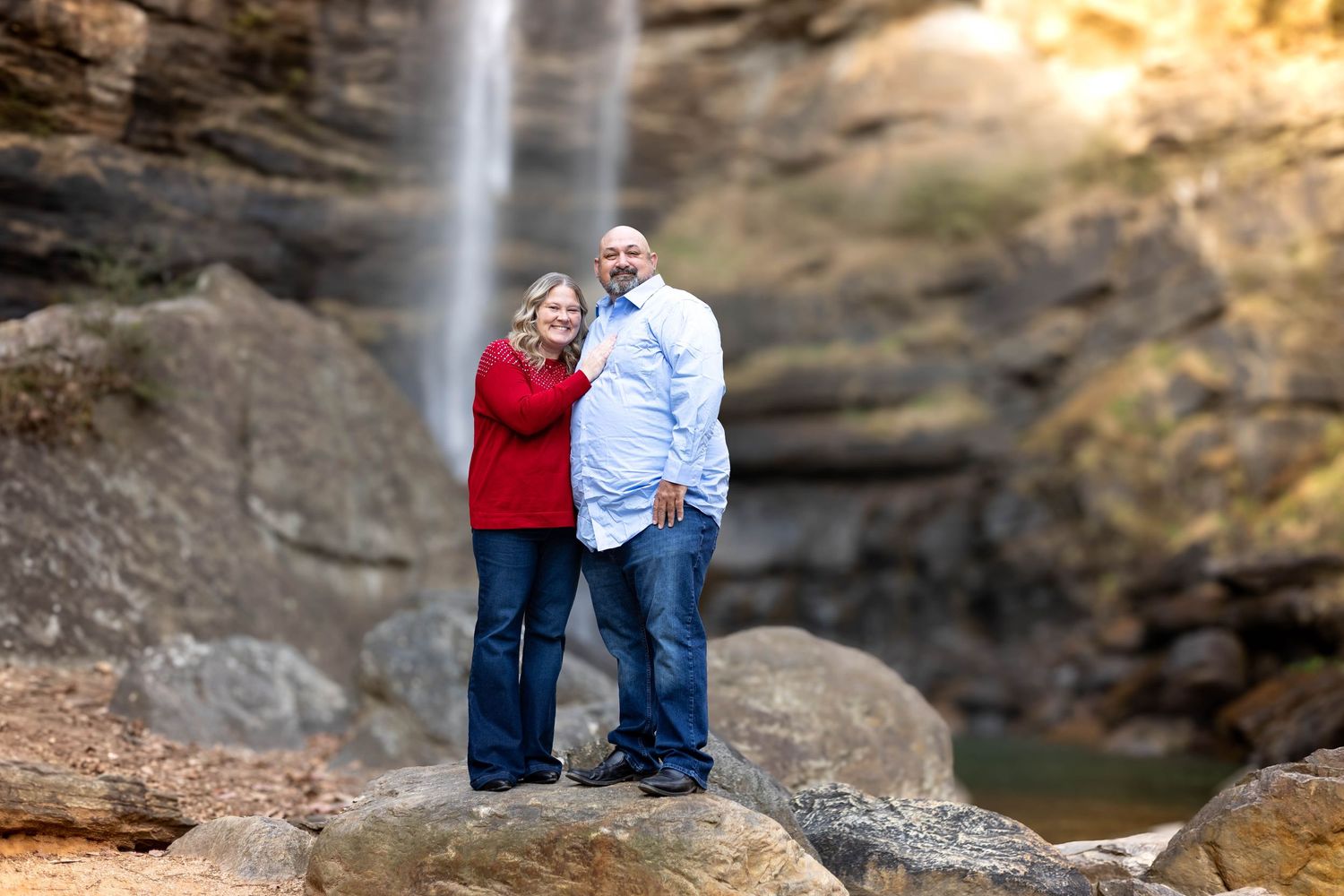 Couple in coordinating red and blue outfits posed on rocky outcrop with waterfall backdrop at Toccoa Falls in northeast Georgia.