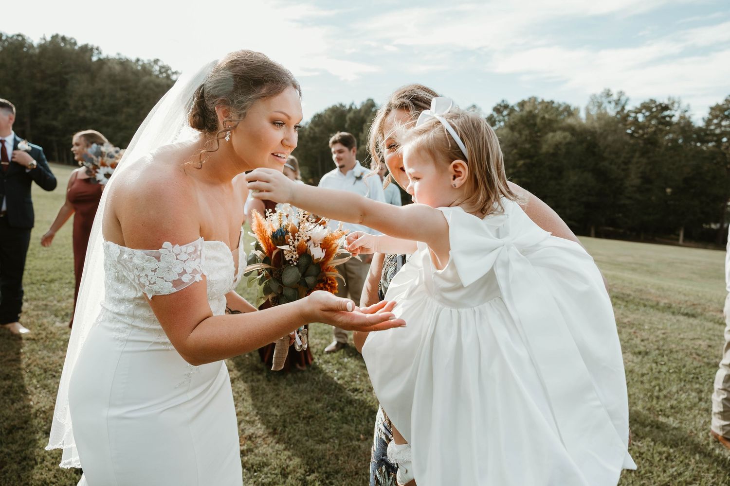 Bride in white wedding dress twirls with flower girl in matching white dress outdoors in grassy field.