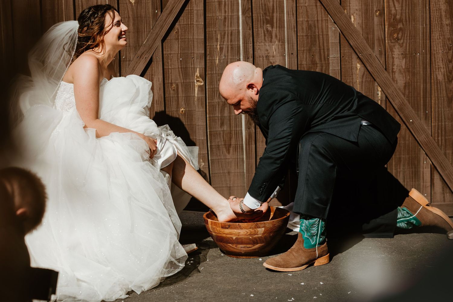 Groom washing bride's feet in wooden bowl during rustic barn wedding ceremony, bride in white gown laughing joyfully.