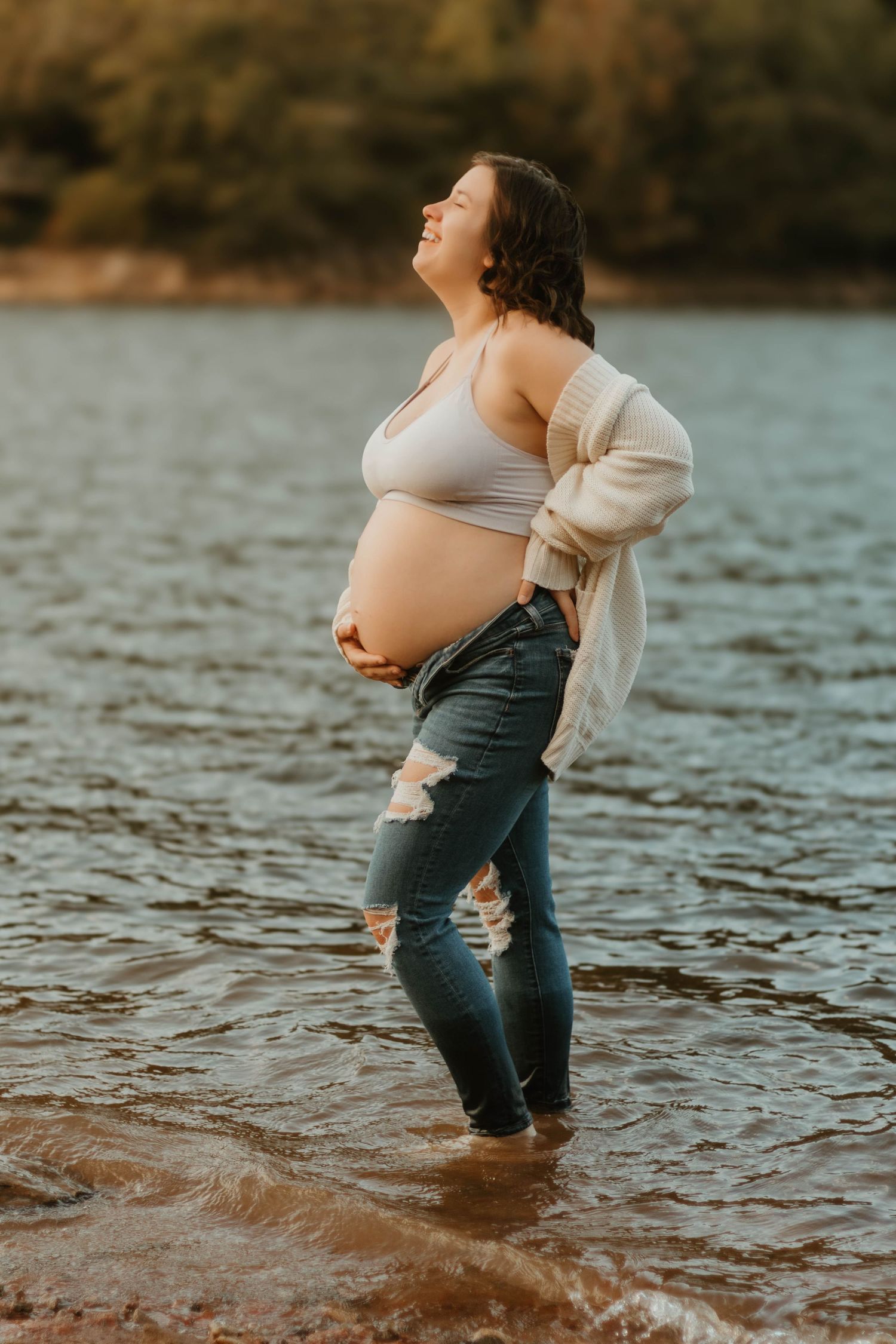 Serene lakeside maternity portrait with expecting mother in ripped jeans and cream cardigan at Lake Hartwell in Georgia.