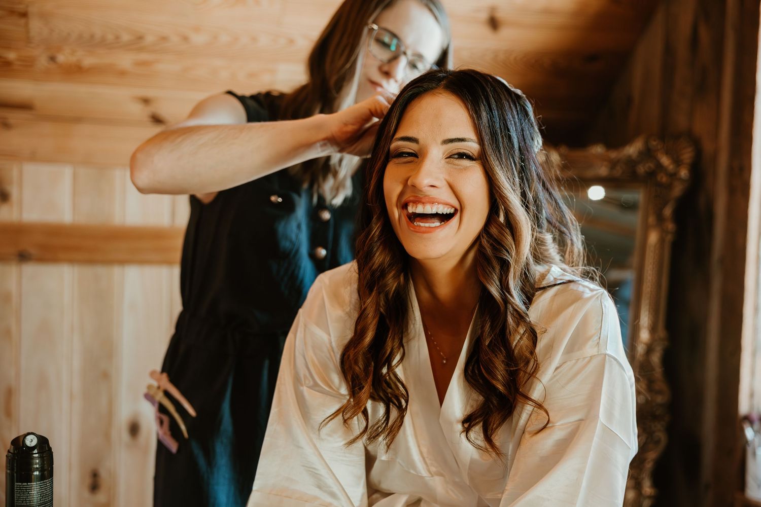 Bride laughing joyfully while hairstylist styles her wavy brown hair in a rustic wooden cabin setting.