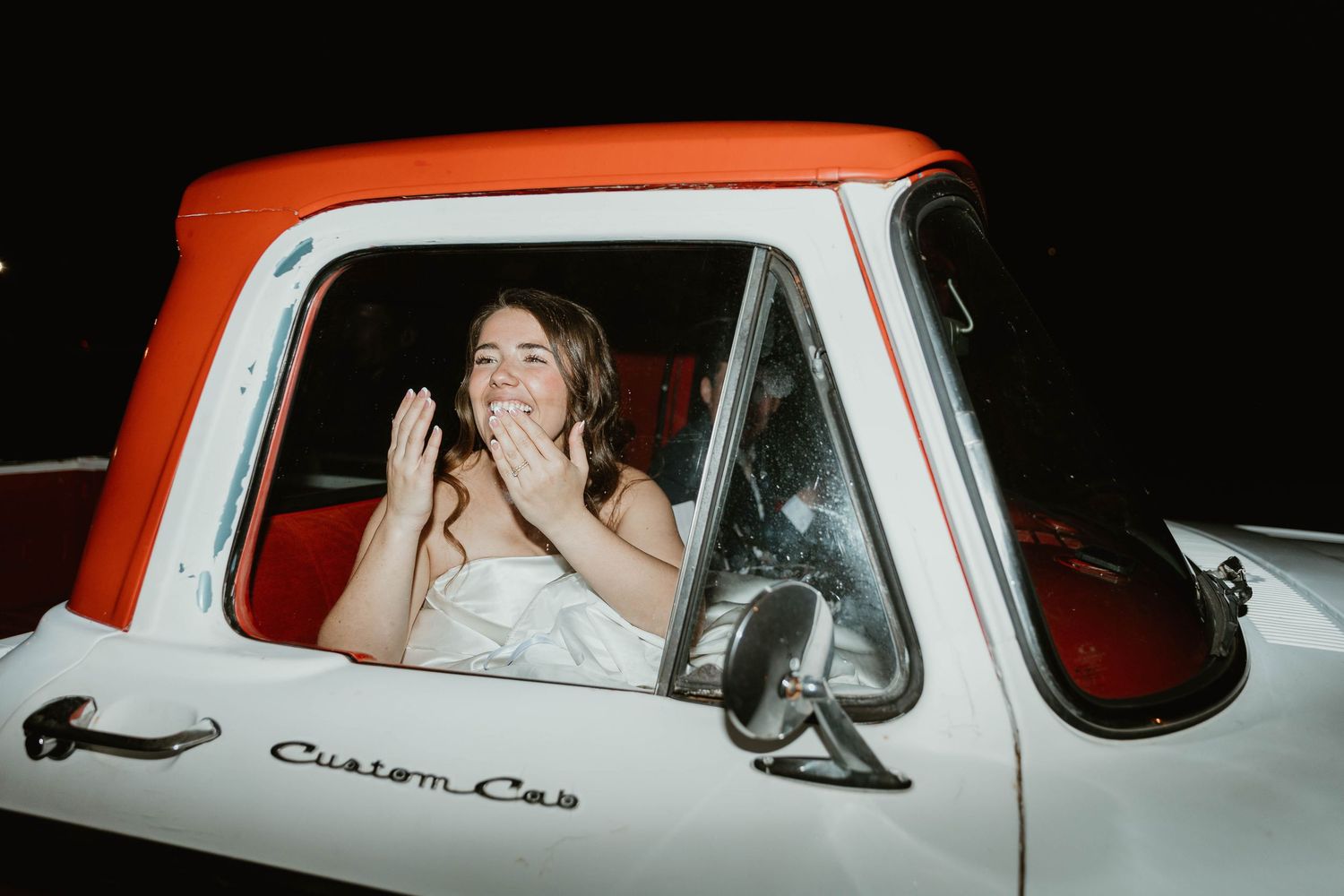 Bride in wedding dress sitting in vintage white and orange Custom Cab truck, clapping with joy at night.