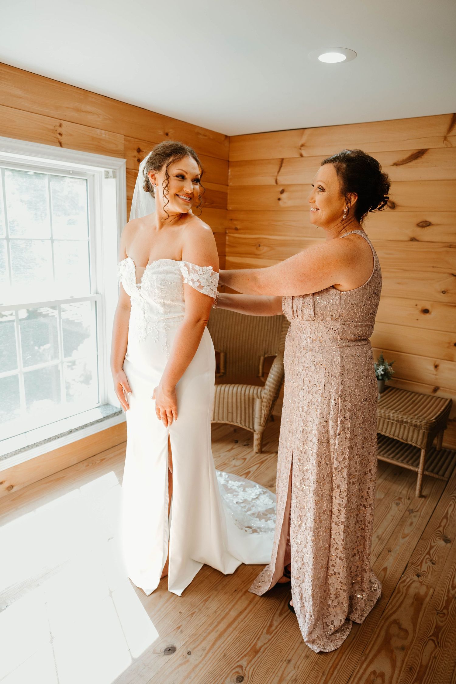 Women in formal dresses prepare for a wedding in a rustic wooden room with natural lighting.