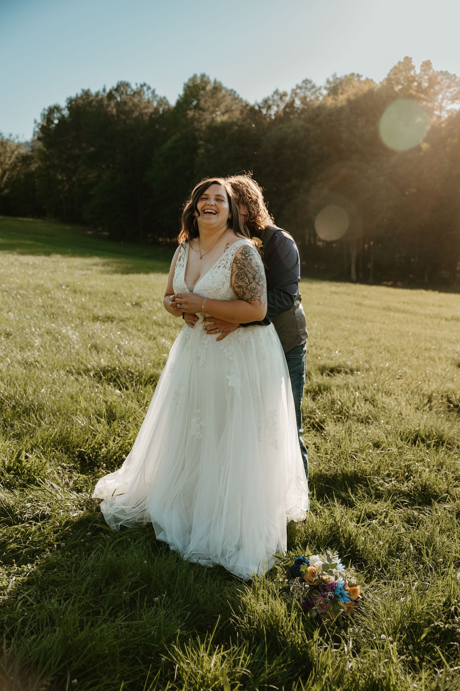 Bride in white lace gown laughing as groom embraces her from behind in a sunlit green field.
