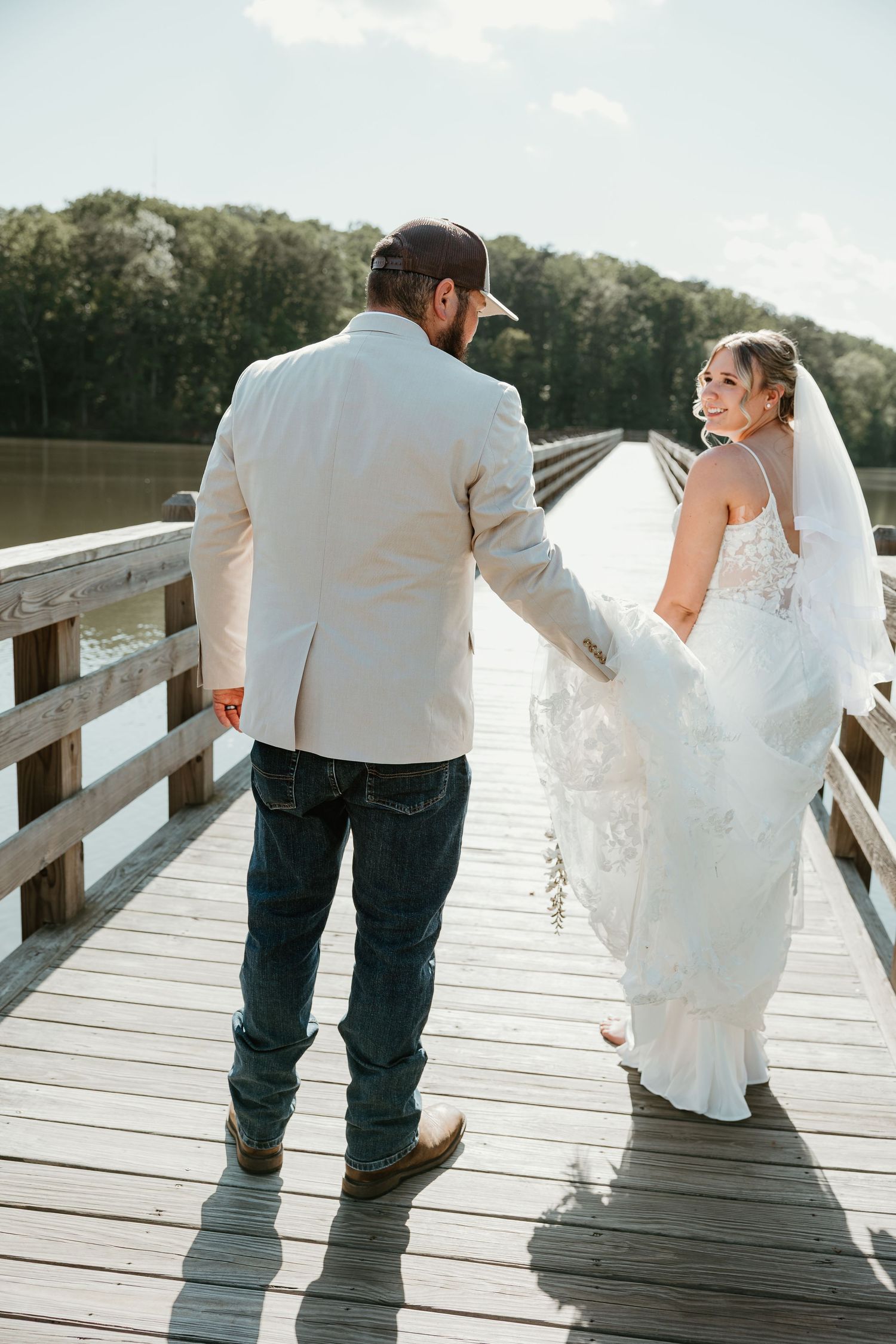 Bride and groom walking hand-in-hand on a wooden pier, bride in white gown glancing back, groom in jeans and blazer.