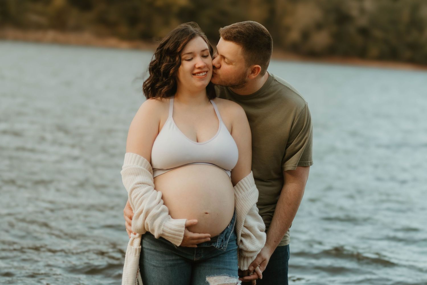 Romantic lakeside maternity photo with couple embracing in casual wear during golden hour at Broken Bridges.