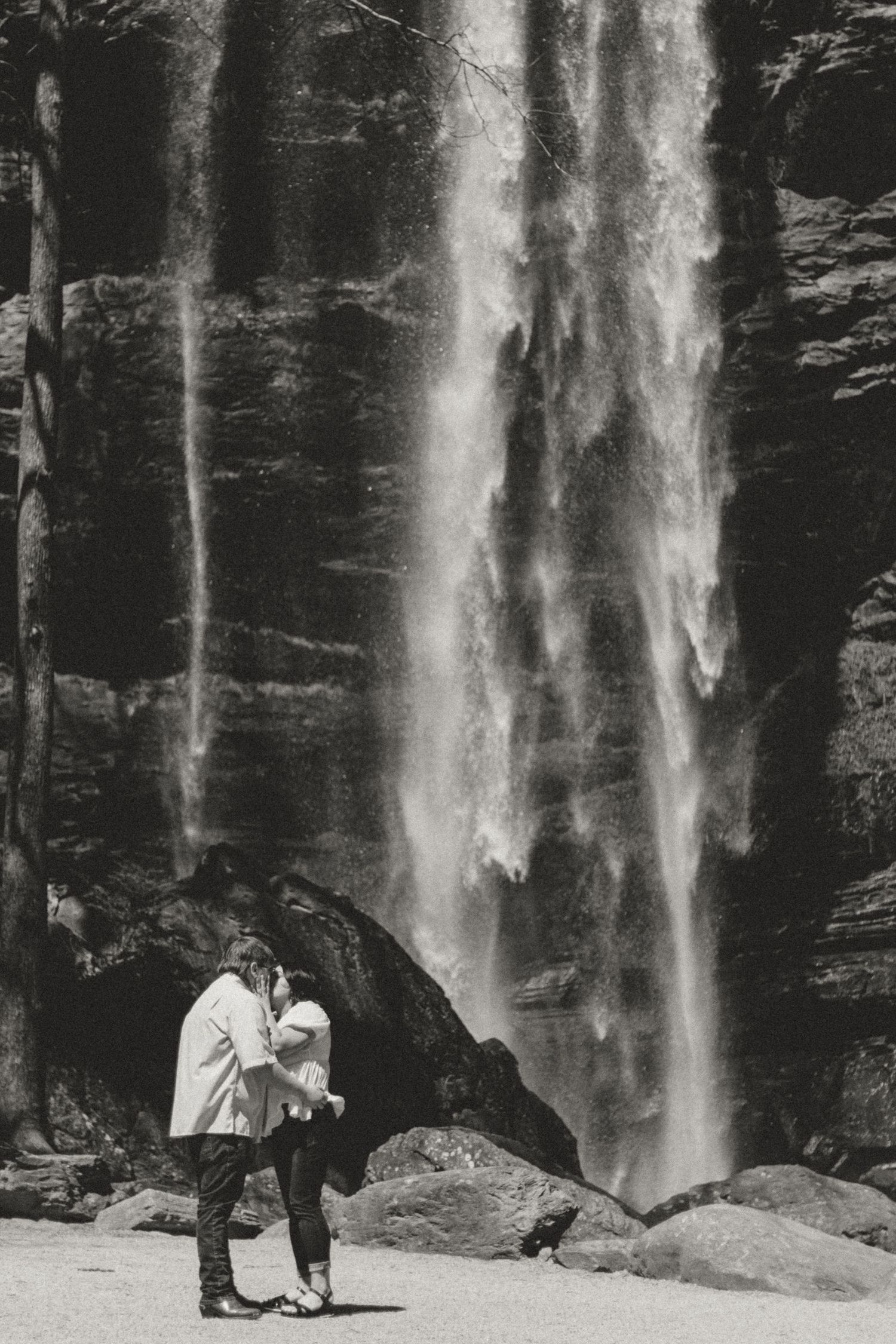 A couple embraces in front Toccoa Falls in northeast Georgia against dark rocky cliffs in black and white.