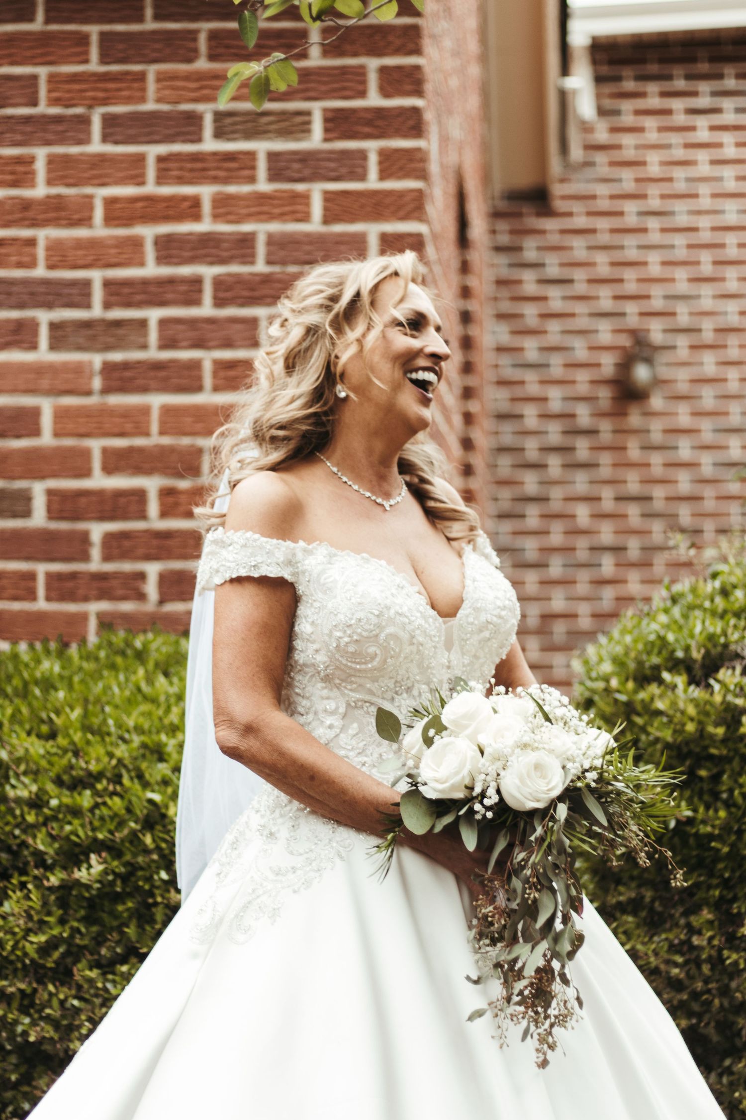 Elegant bride in white off-shoulder gown smiles outdoors against red brick wall holding white floral bouquet.
