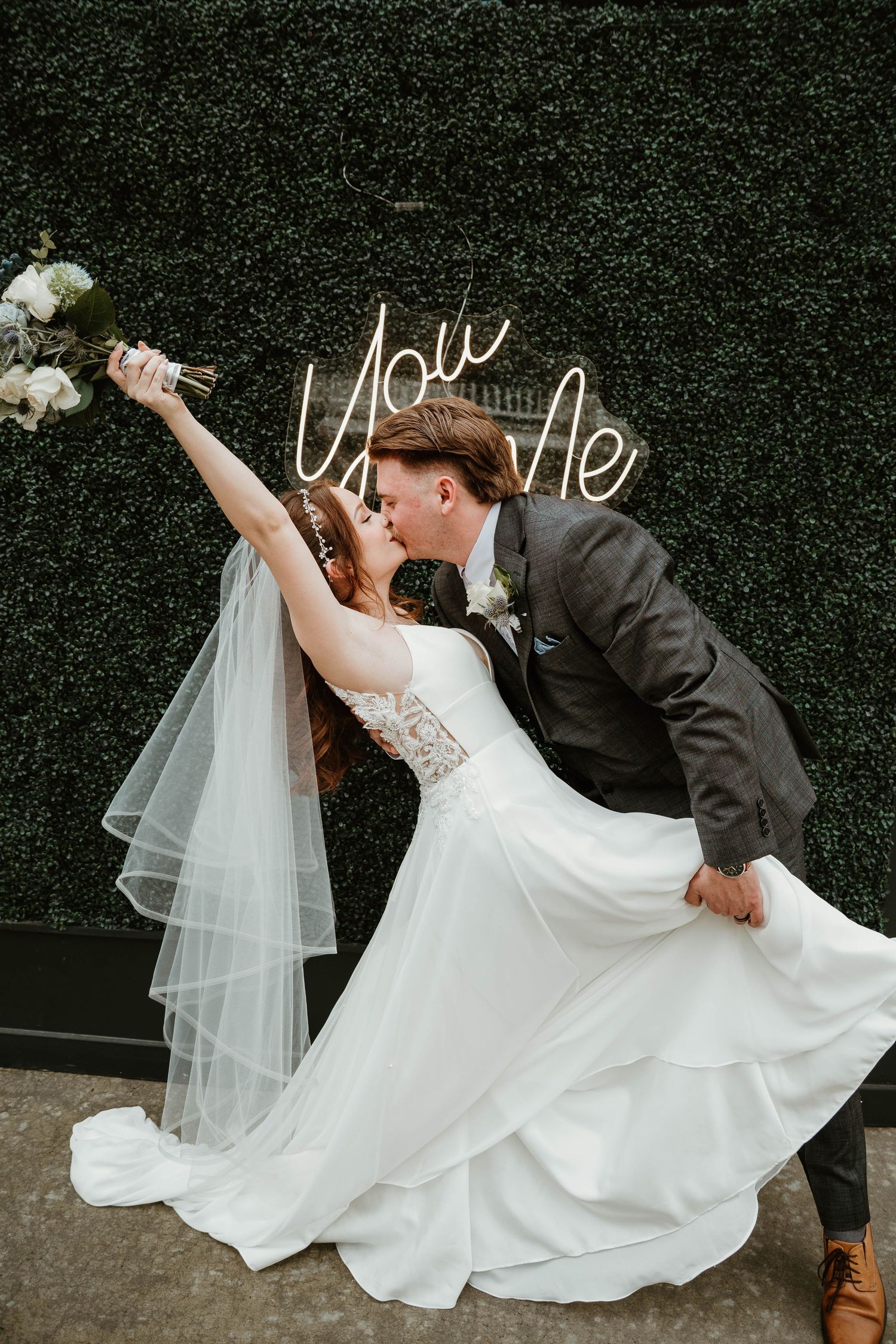 Bride and groom share a romantic dip kiss in front of a green boxwood wall with a glowing neon sign.