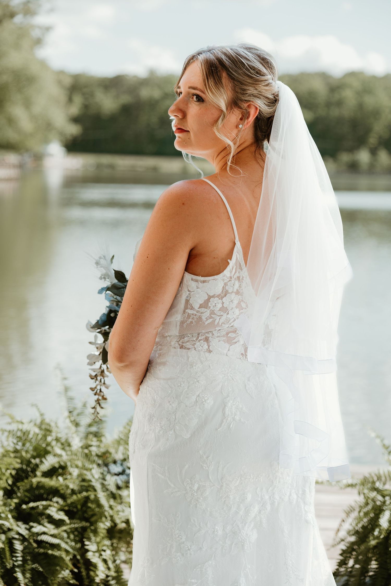 Bride in lace wedding dress and veil holds bouquet by lakeside, looking over shoulder in outdoor setting.