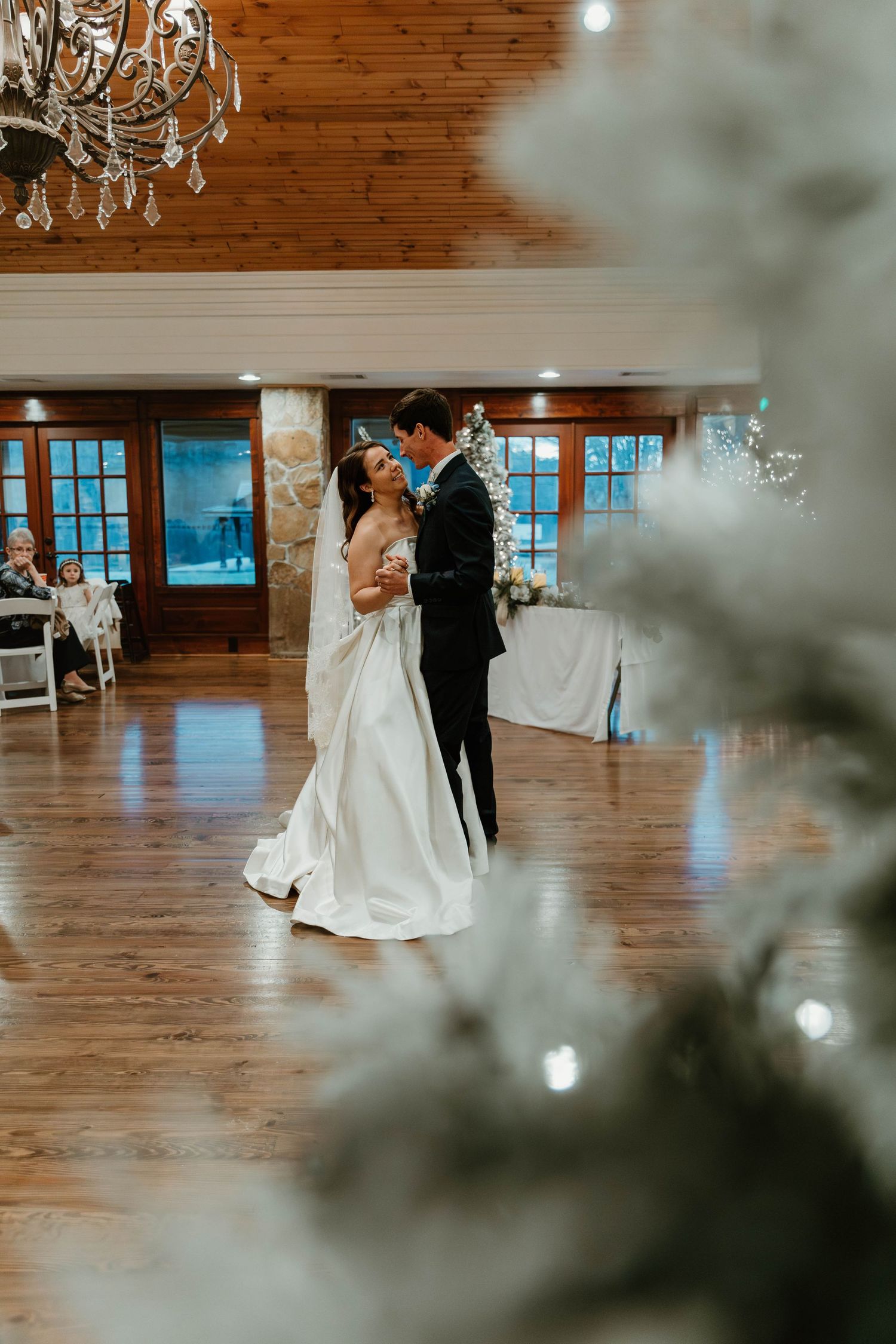 A wedding reception venue features wood paneling, chandeliers, and couples dancing on a polished floor.
