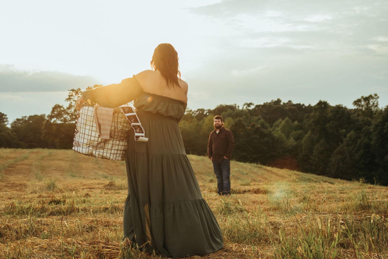 Expecting couple in a golden field at sunset with long dress and vintage-style props creating a dreamy maternity photo in northeast Georgia.