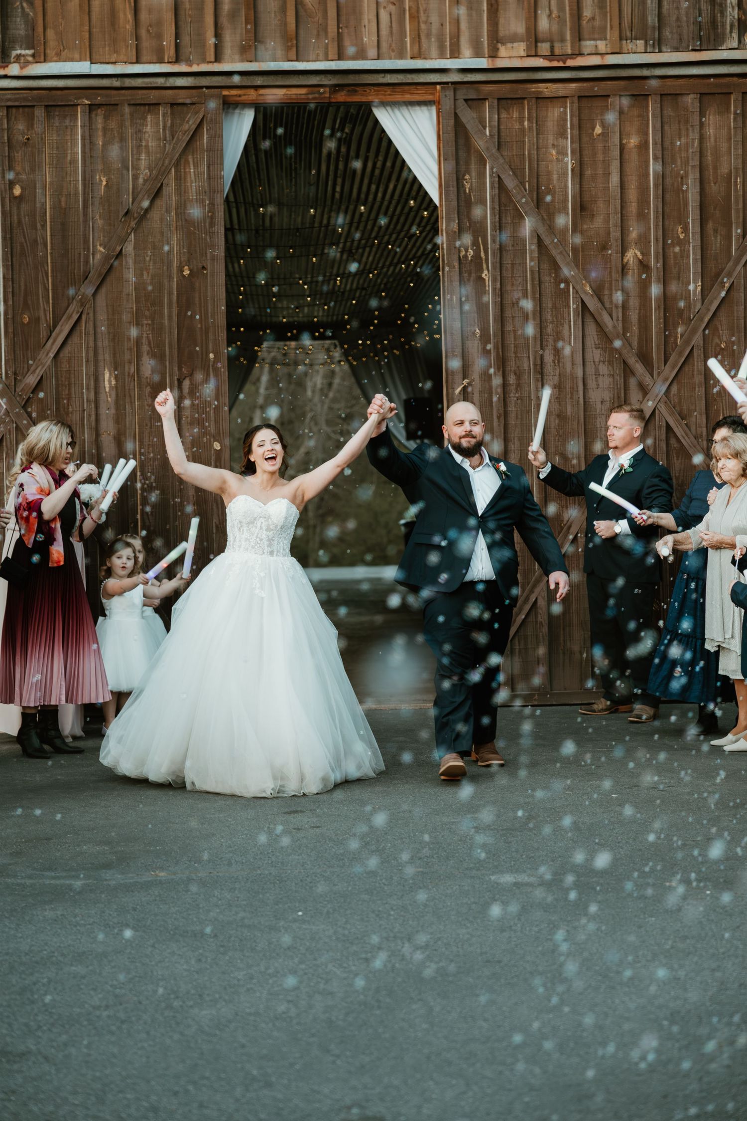 Joyful bride and groom exit rustic barn doors amid confetti shower, raising hands in celebration with wedding guests.