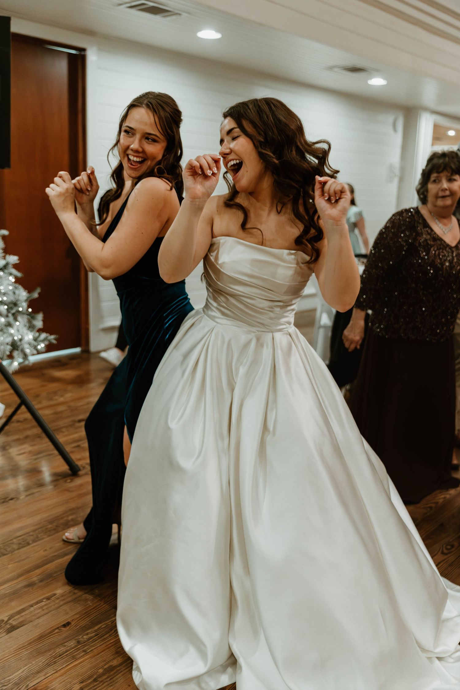 Bride in white ballgown dancing joyfully with bridesmaid in teal dress at wedding reception.