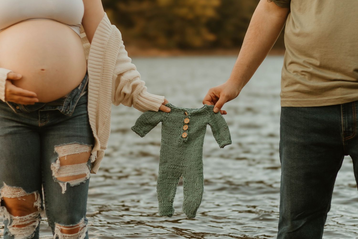 Couple holds tiny teal baby outfit by the water during maternity photoshoot near Lake Hartwell.