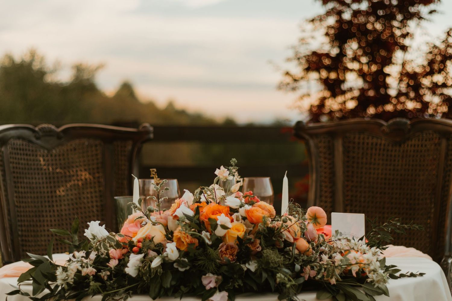 A sunset wedding reception setup features elegant floral centerpieces on rustic wooden tables with glowing candles.
