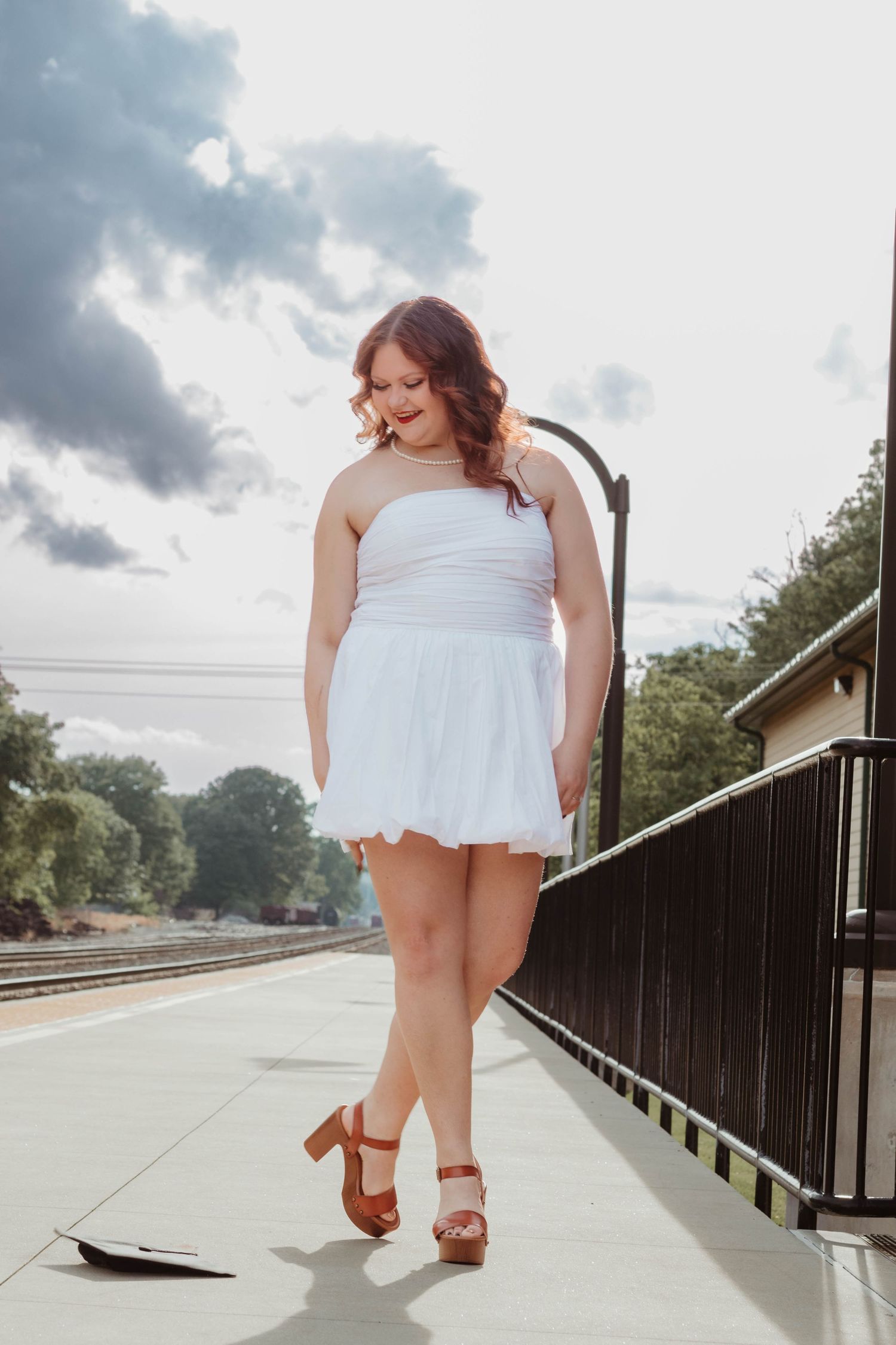 A person in a white summer dress and brown sandals poses on a train platform on a cloudy day in Toccoa, Georgia.