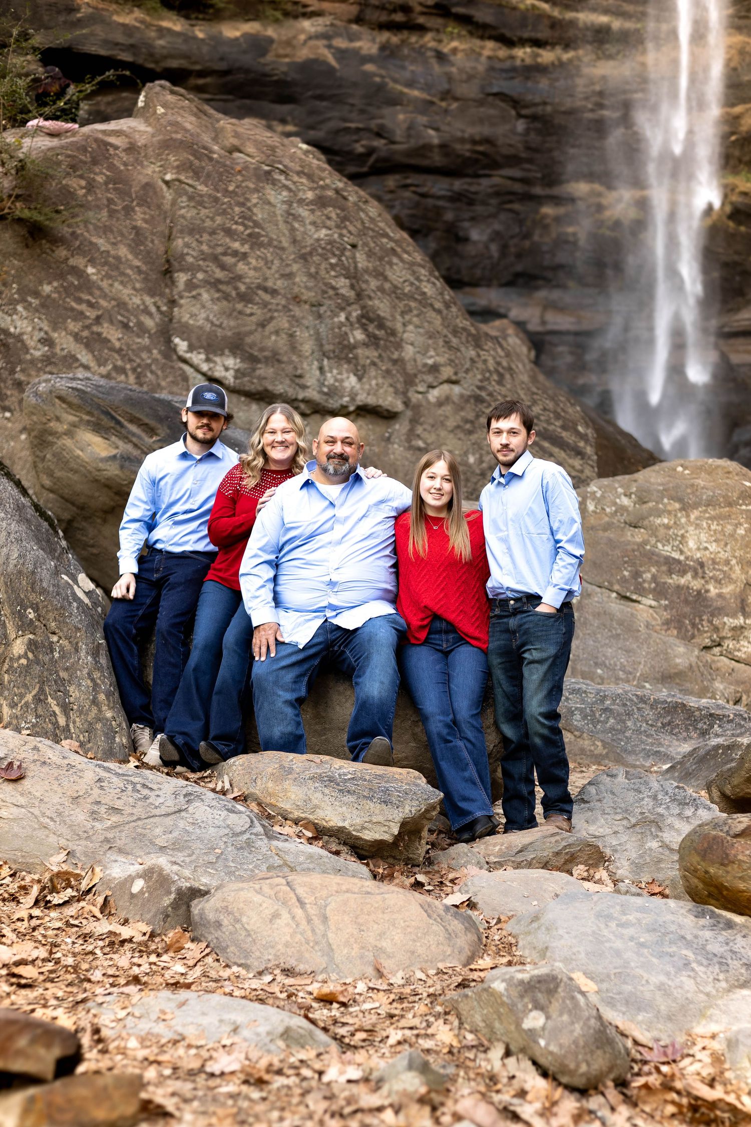 Family group poses on rocky terrain with dramatic waterfall backdrop in coordinating red and blue outfits at Toccoa Falls.