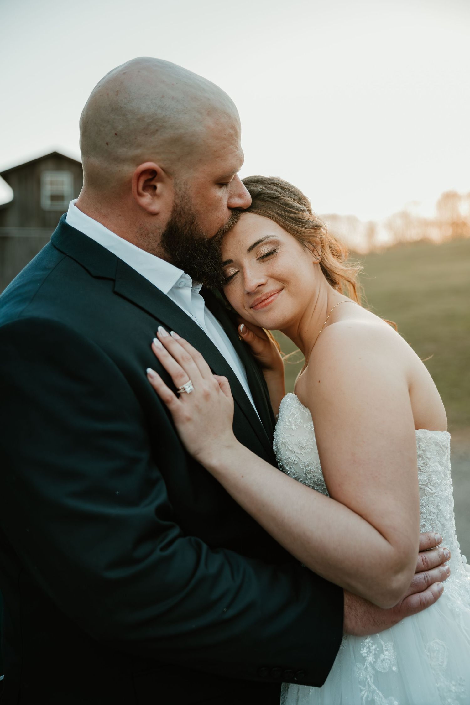 Groom in dark suit kisses brides temple as she smiles peacefully during golden hour portrait session.
