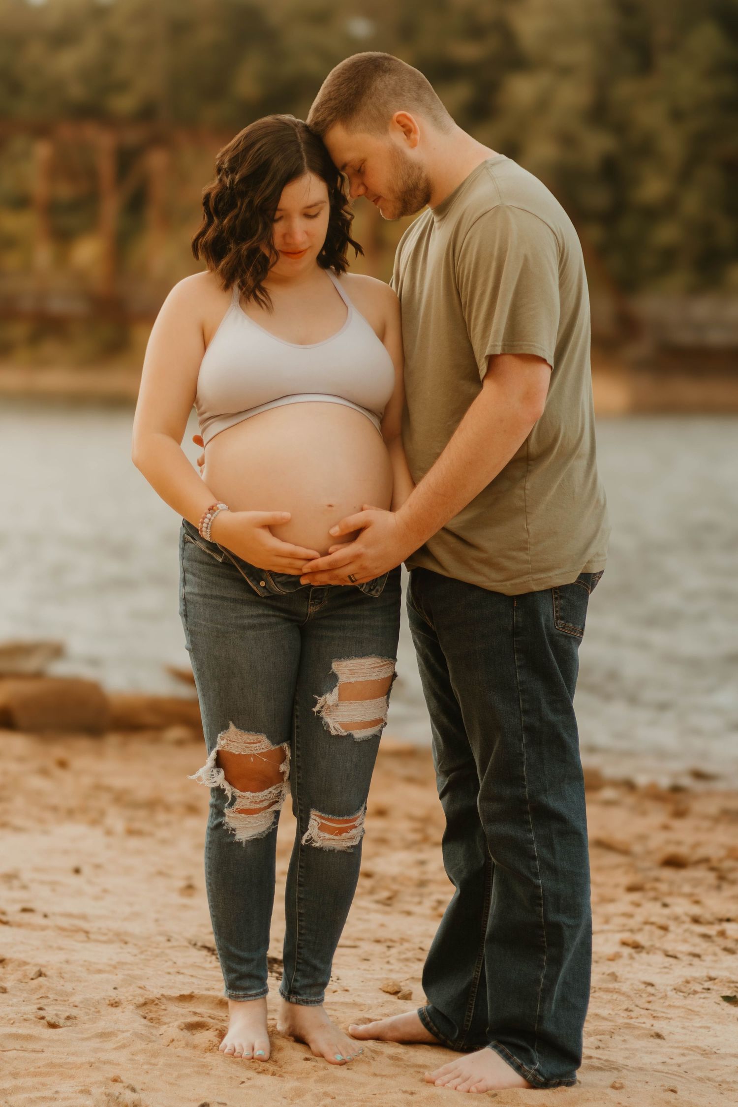 Tender lakeside maternity moment with couple in casual wear touching pregnant belly at Broken Bridges in Toccoa, GA.