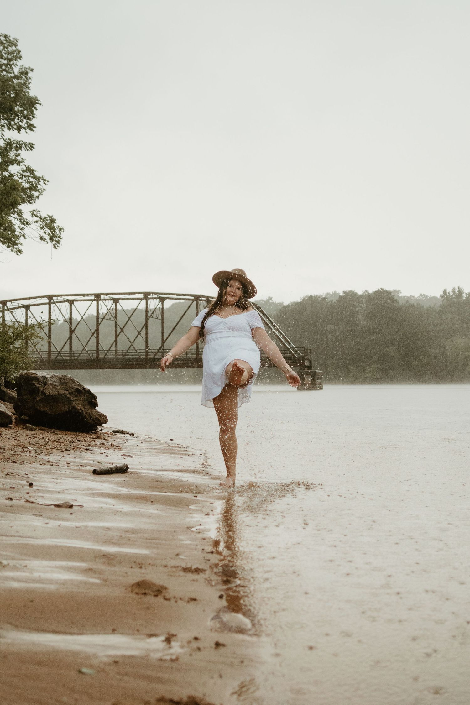 Person in a flowing dress playfully kicks water at the lake's edge with a brown hat on Lake Hartwell.