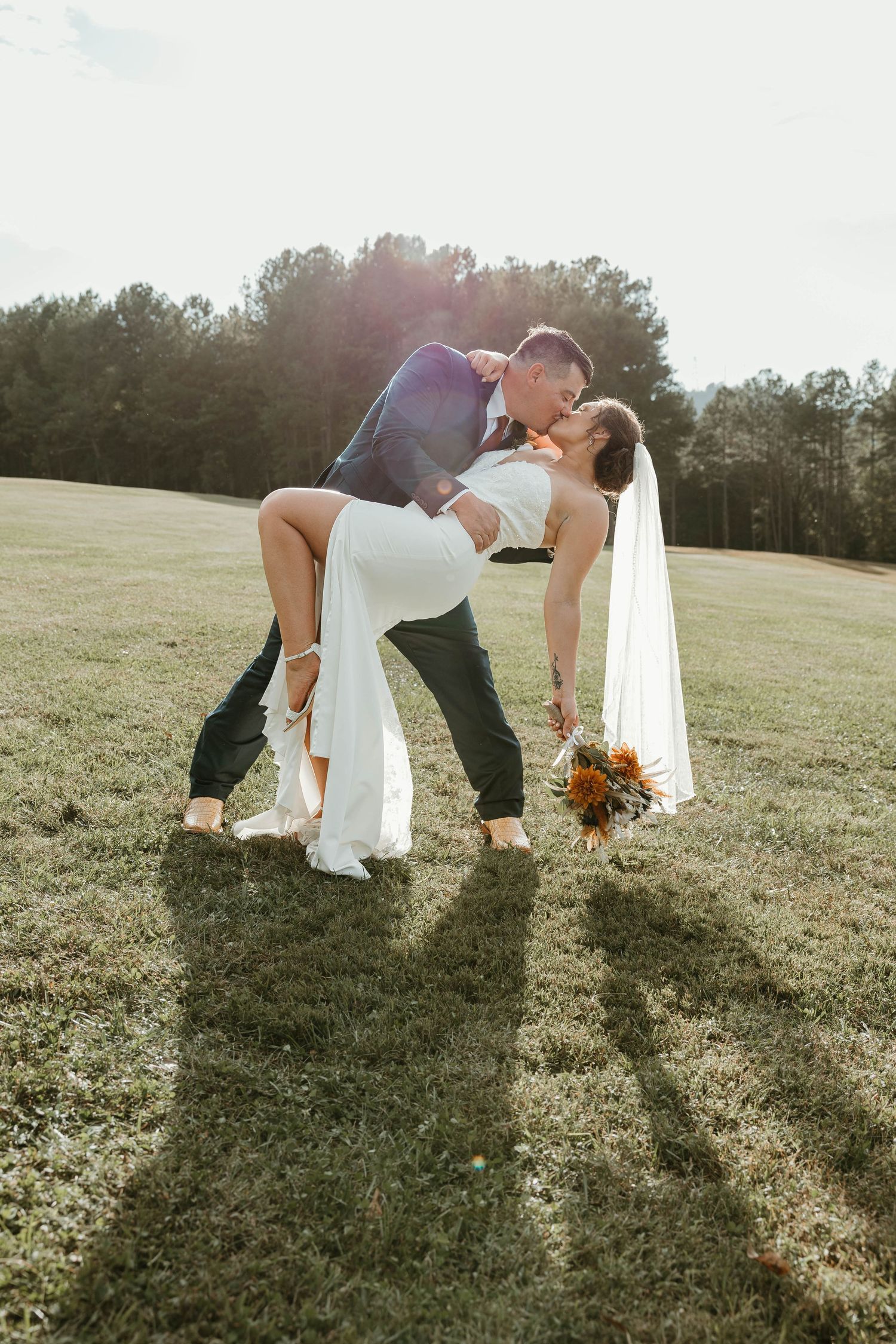 A romantic wedding photo of a couple sharing a kiss in a sunlit field while holding a bouquet of orange flowers.