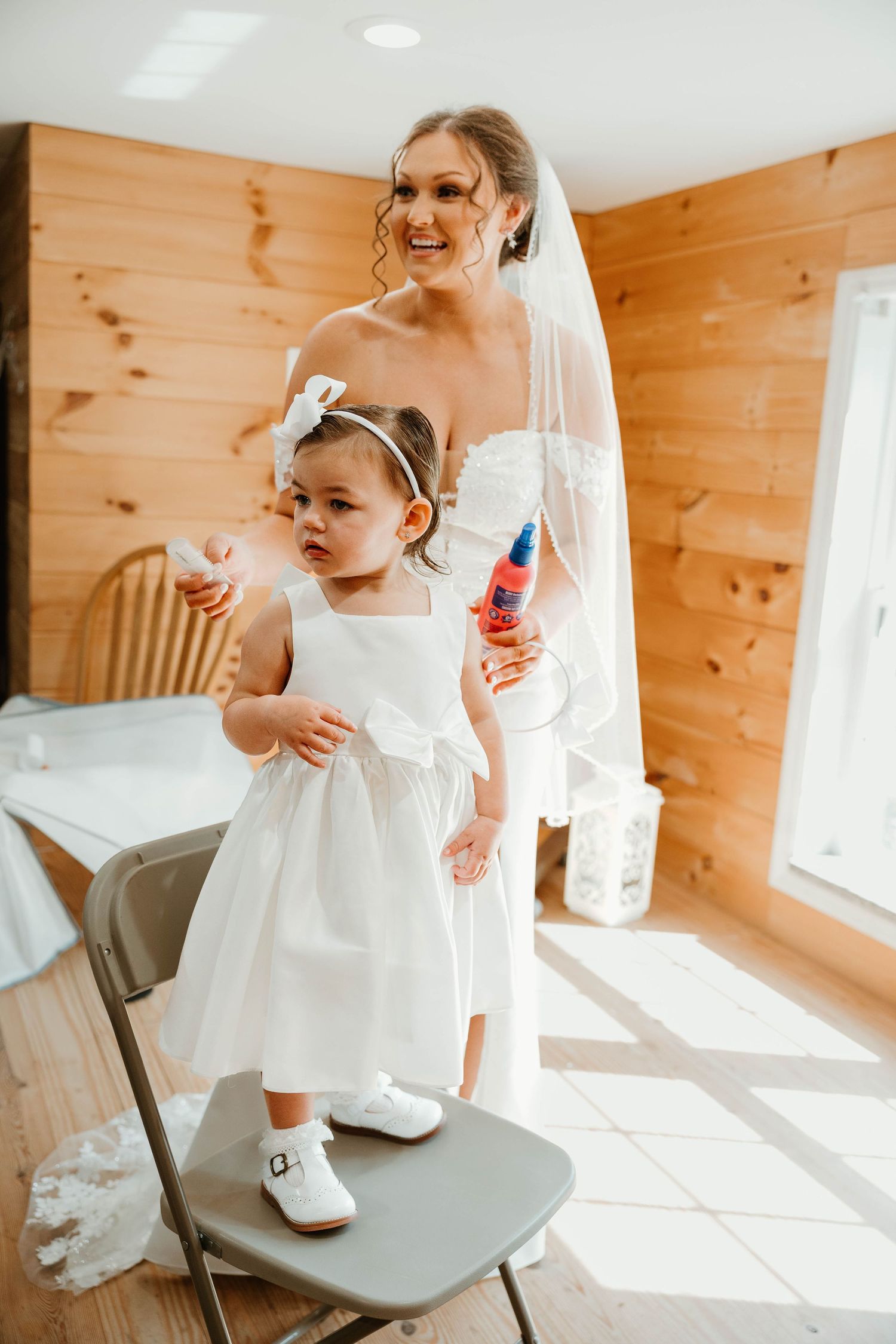 A small child in a white dress and shoes stands on a gray chair in a wooden-paneled room.