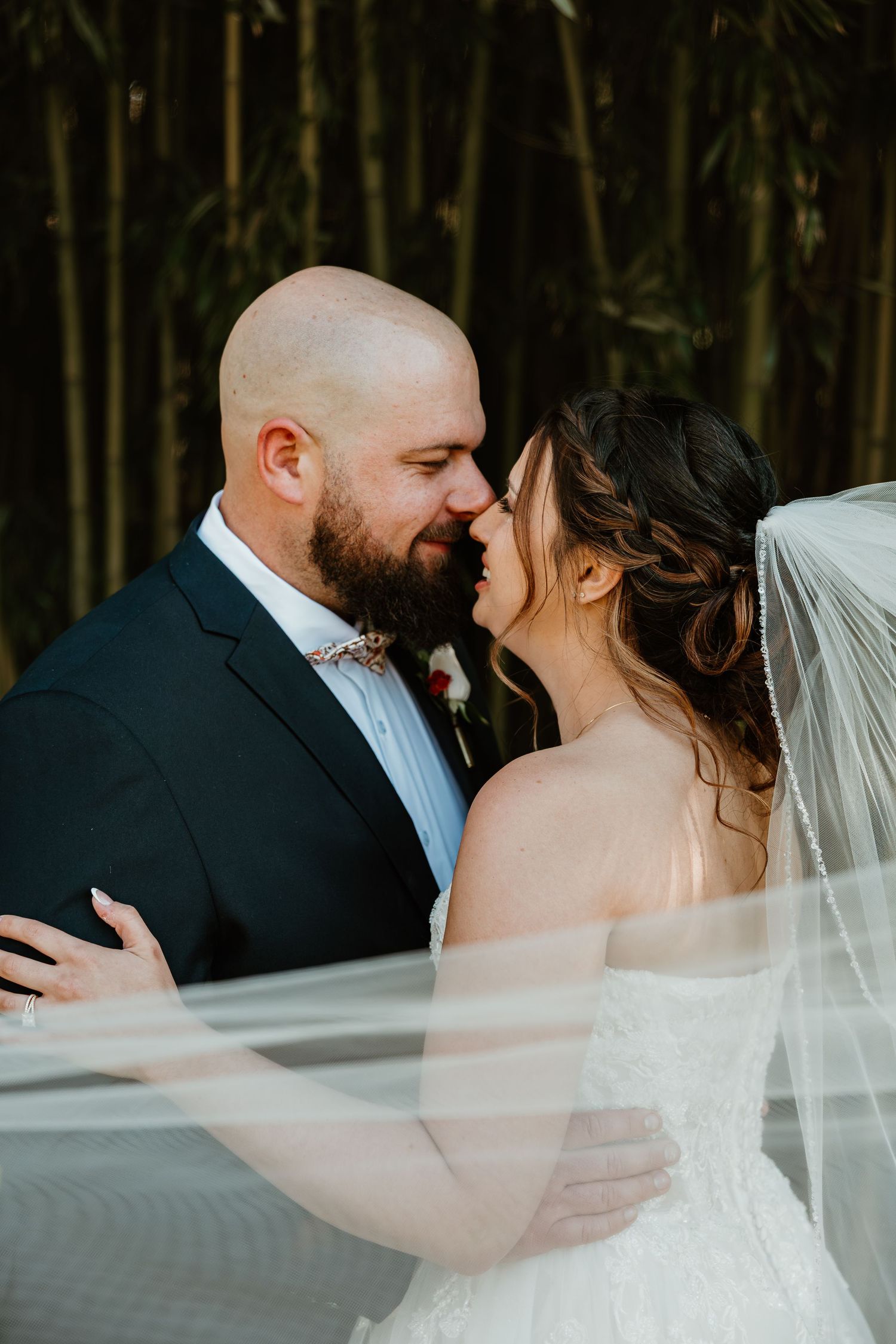 Bride and groom sharing a romantic almost-kiss moment, with veil flowing, bamboo backdrop, groom in navy suit.