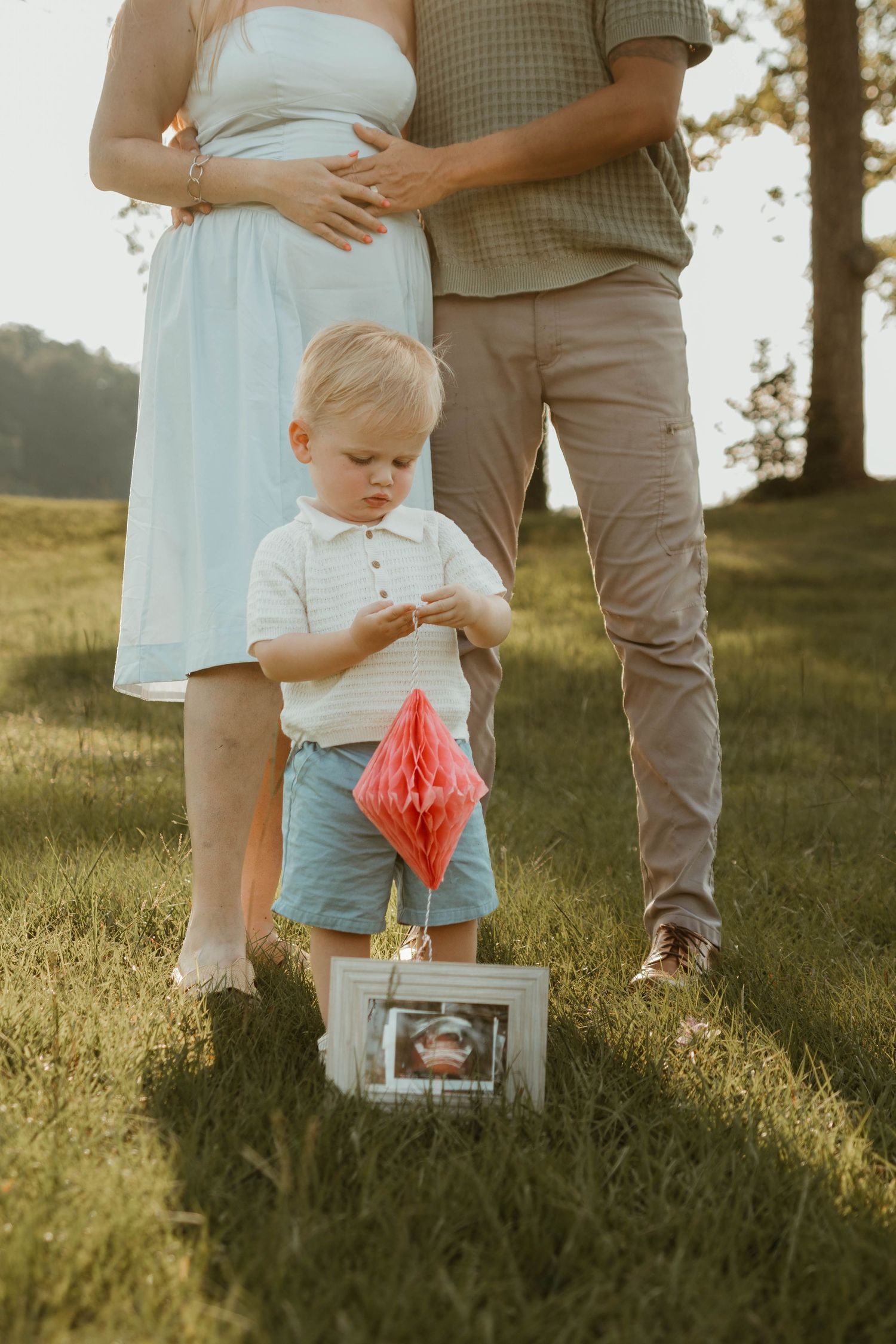 A toddler examines a pink honeycomb decoration while standing in a park during golden hour for a pregnancy announcement in northeast GA.