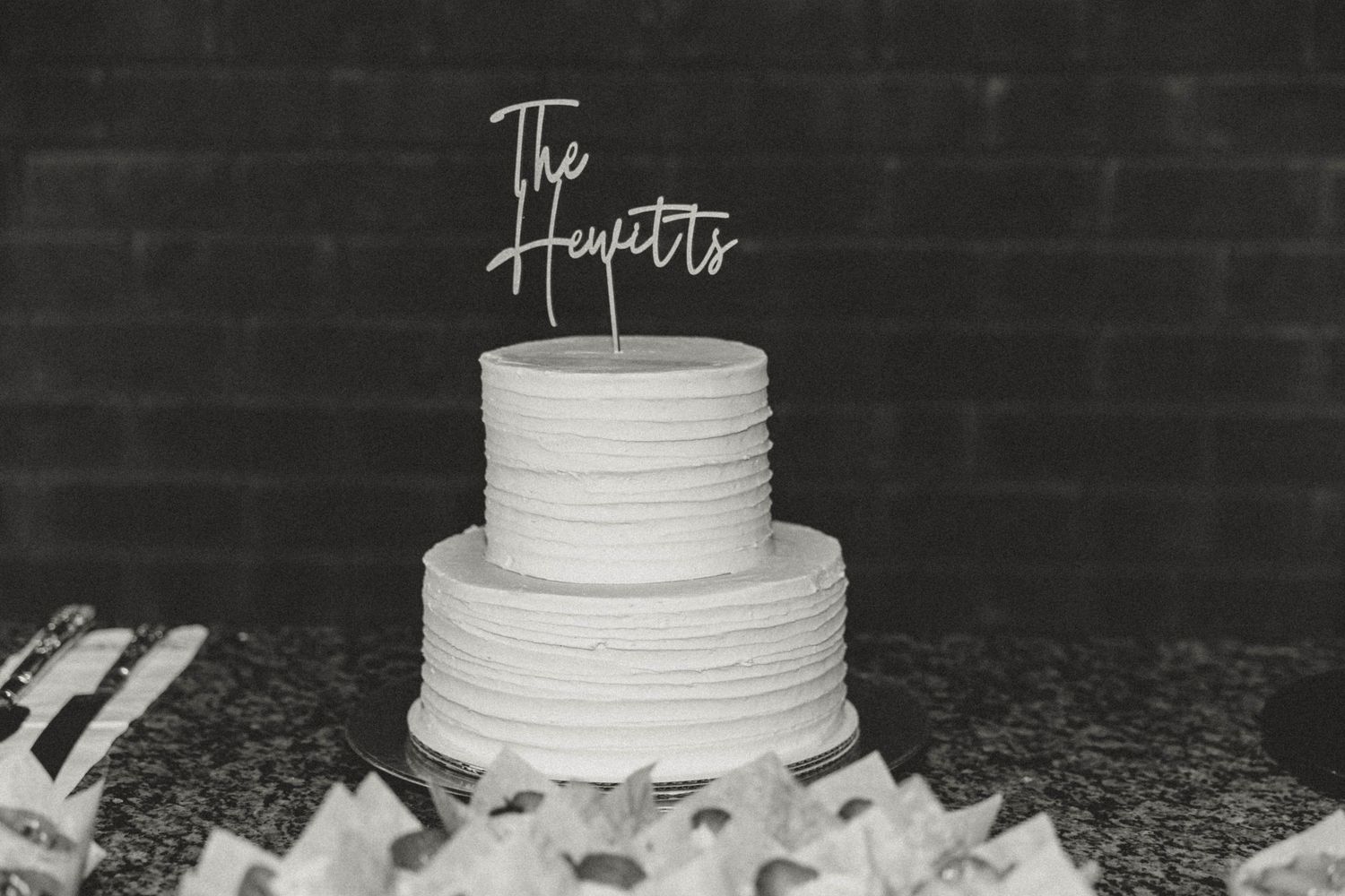 A close-up view of a simple white tiered wedding cake against a dark background.