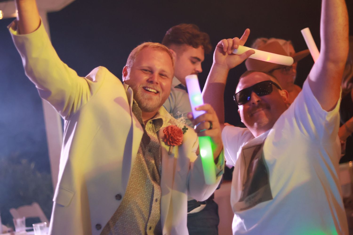 A group of groomsmen in white outfits dancing and singing together at a wedding reception with colorful lighting at Cherry Ranch in Toccoa.