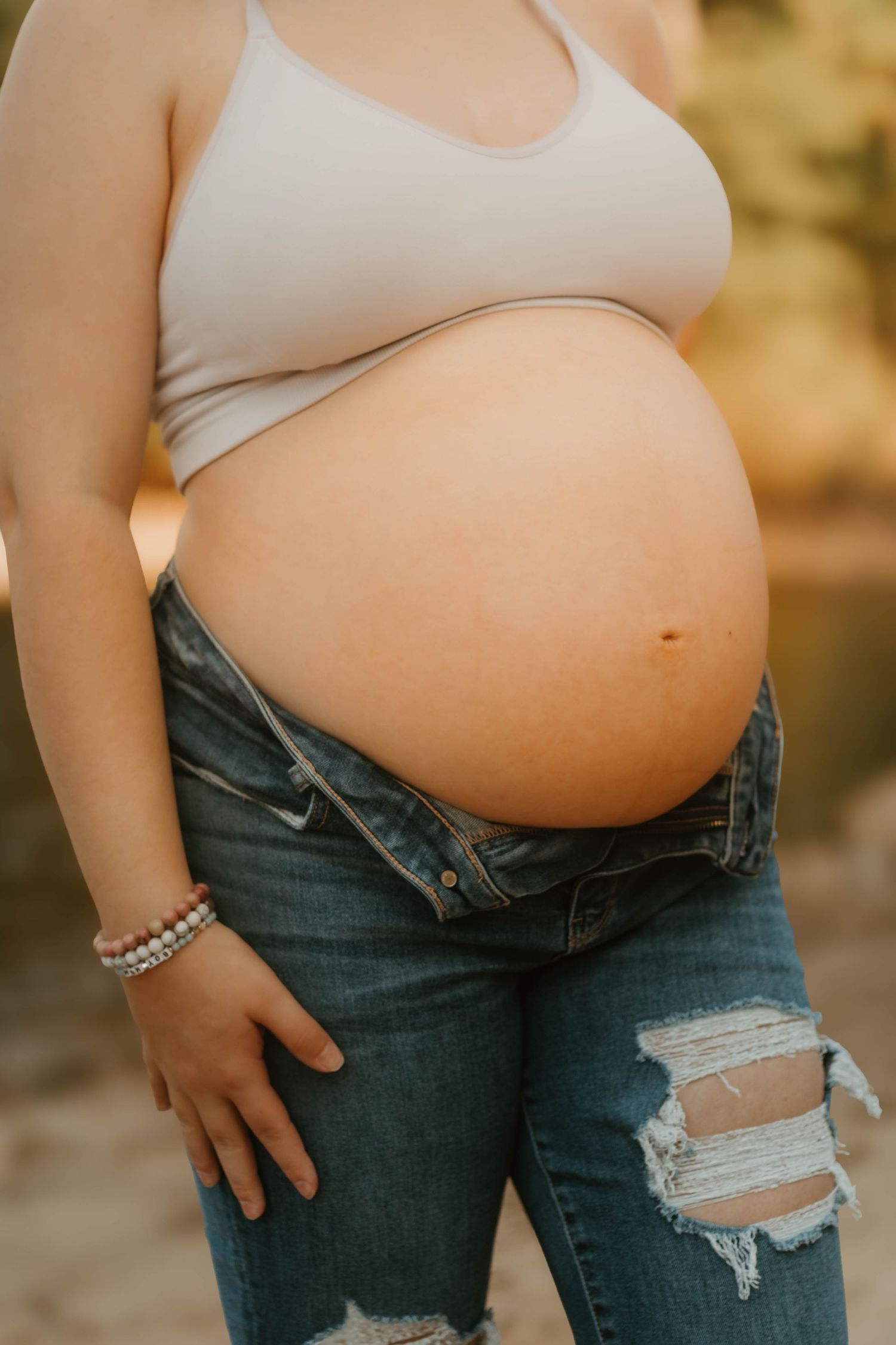 A close-up of a pregnant belly in ripped jeans and white crop top against golden light on Lake Hartwell.