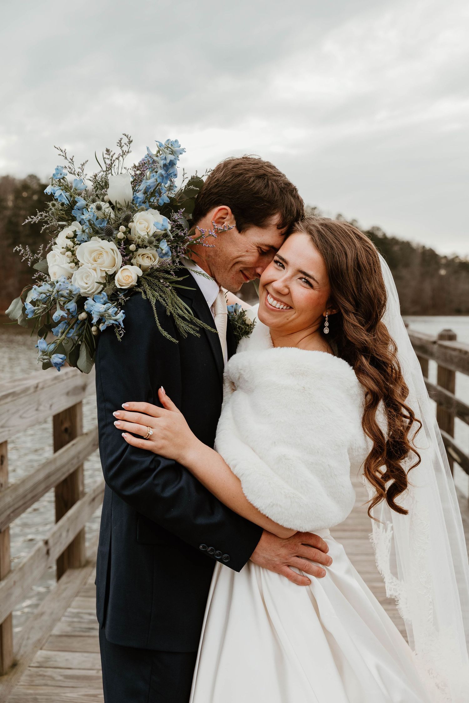 Bride and groom embracing on a wooden dock, bride holding blue and white bouquet, wearing white gown with fur wrap.