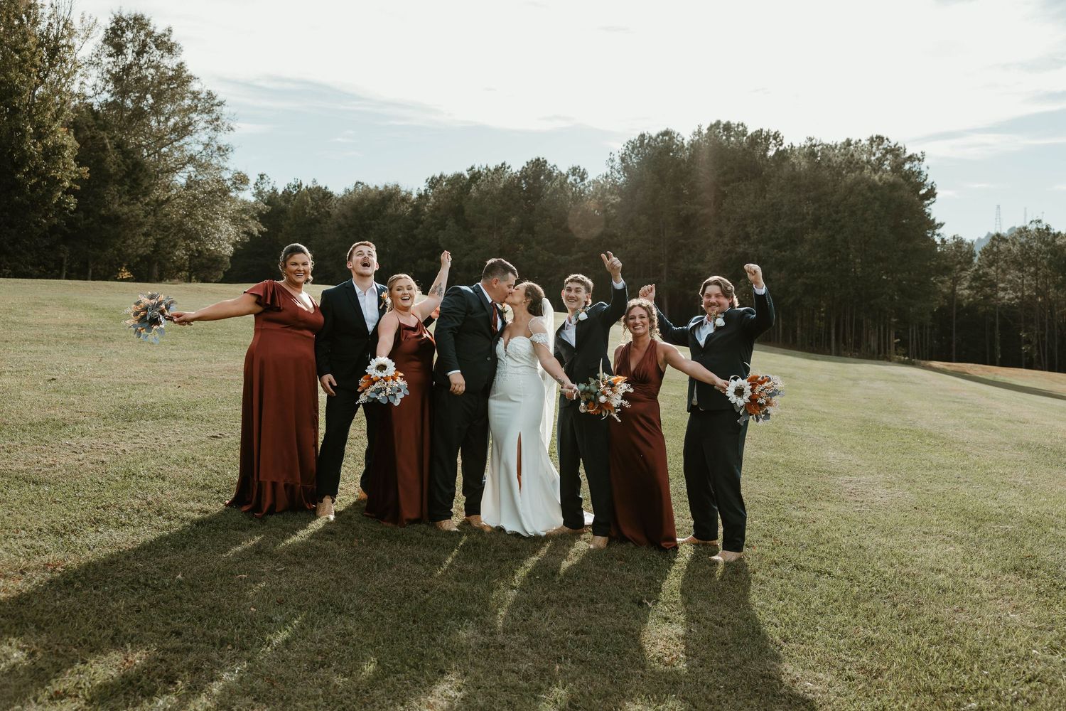Wedding party members celebrate joyfully while posing together in a scenic outdoor field at sunset.
