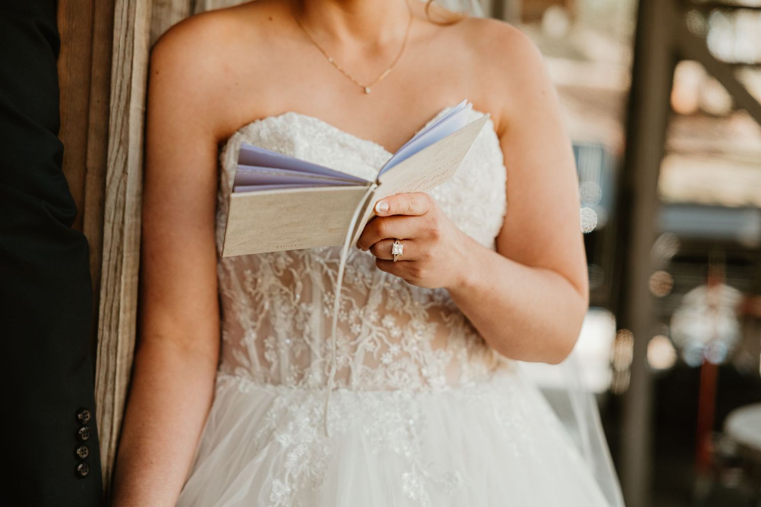 Bride in strapless lace wedding gown holding vow booklets at rustic wedding venue.
