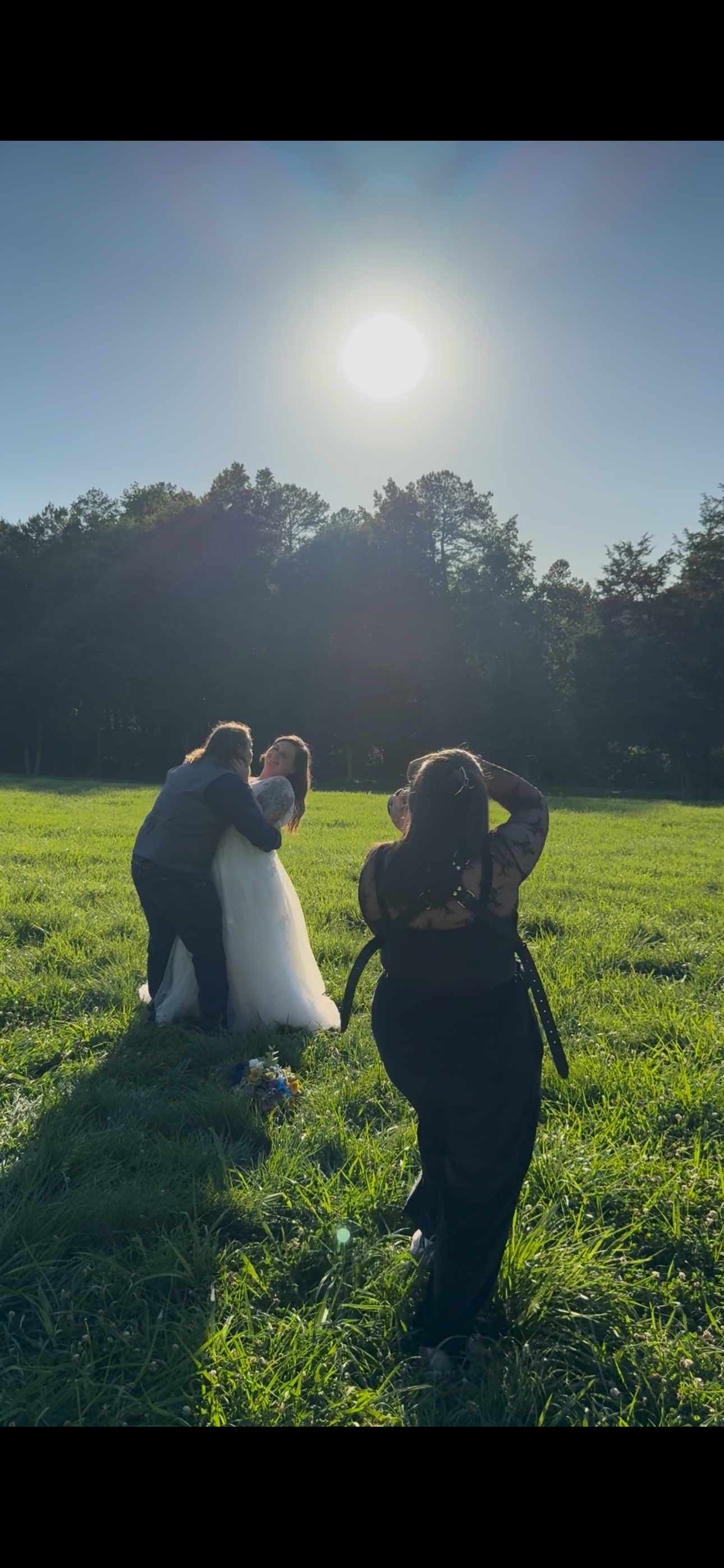 Photographer capturing bride and groom kissing in sunny green field with trees in background during outdoor wedding photo session.