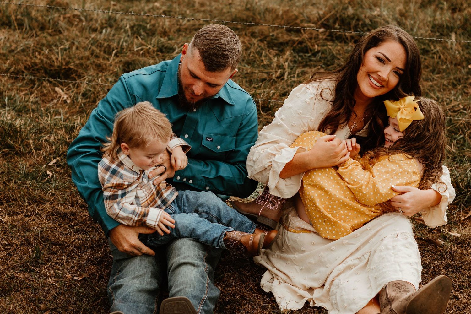 A young family sits together outdoors on brown grass during a fall photoshoot wearing coordinated blue and yellow outfits.