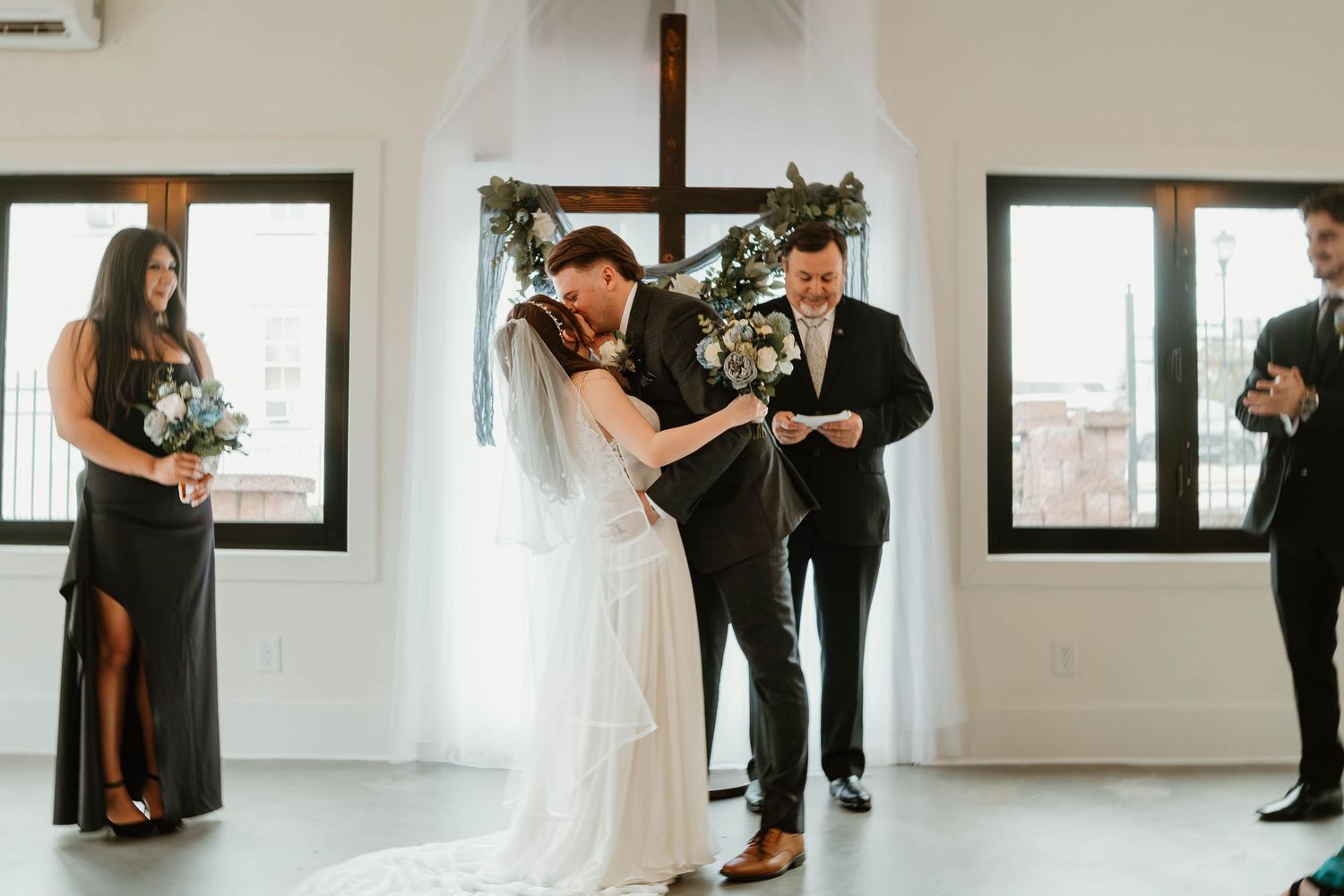 Bride and groom exchange rings at indoor wedding ceremony with cross backdrop, bridesmaid and officiant standing nearby.
