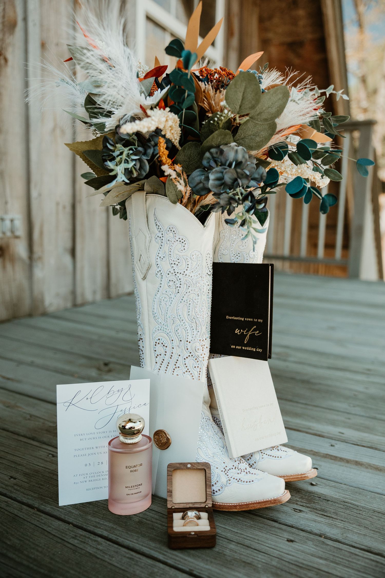 White cowboy boots holding a rustic wedding bouquet with rings, perfume, invitation, and vow book on wooden deck.
