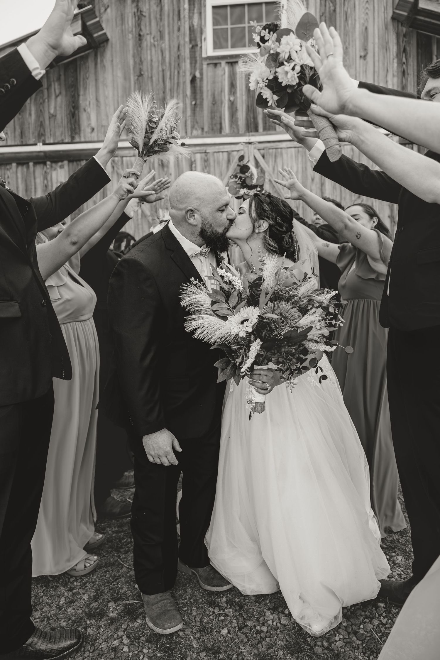 Black and white photo of bride and groom kissing under an arch of raised hands and bouquets outside a rustic barn.
