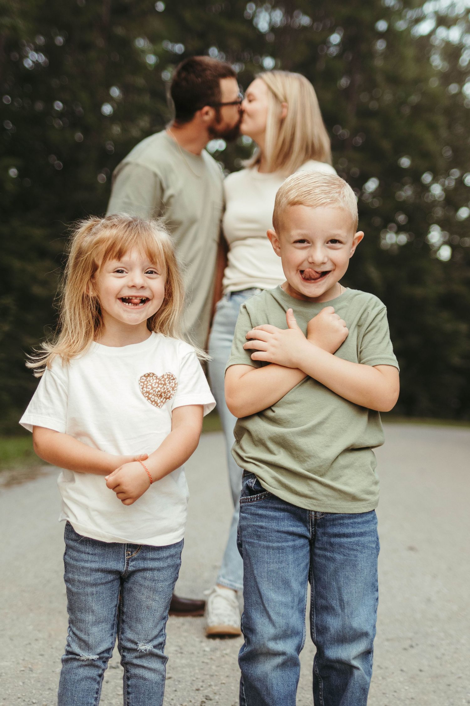 Two children in casual clothing laugh joyfully while standing on a path with adults embracing in the background.