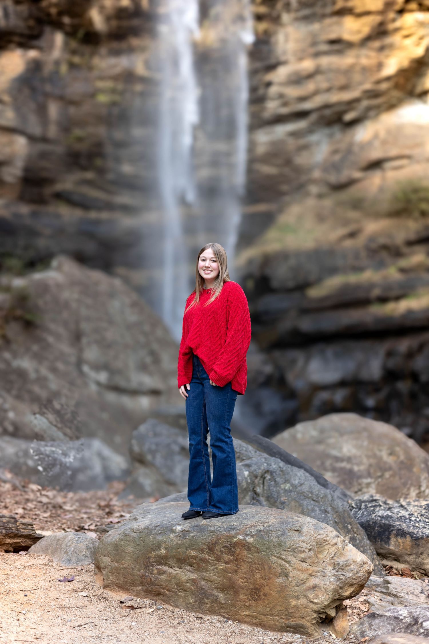 Red sweater and jeans outfit photographed on rocky terrain with majestic waterfall in background at Toccoa Falls in northeast Georgia.
