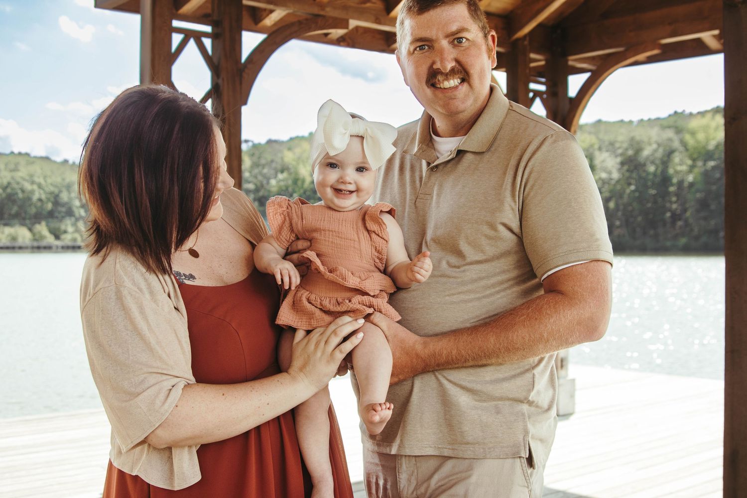 A family wearing earth-toned clothing poses together for outdoor photos by a lake on a sunny autumn day.