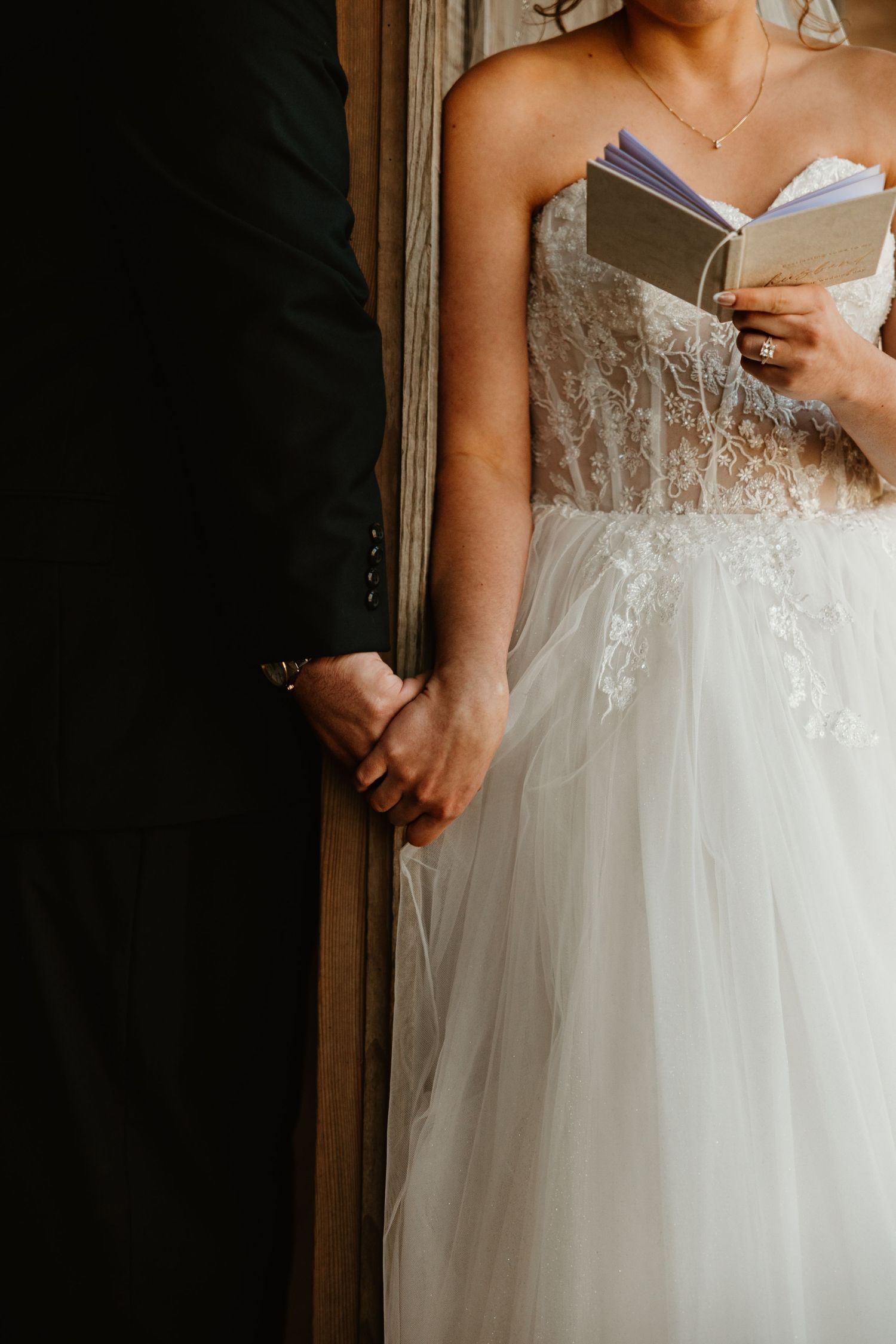 Bride in lace tulle wedding gown holds vows booklet while holding hands with groom in black suit.