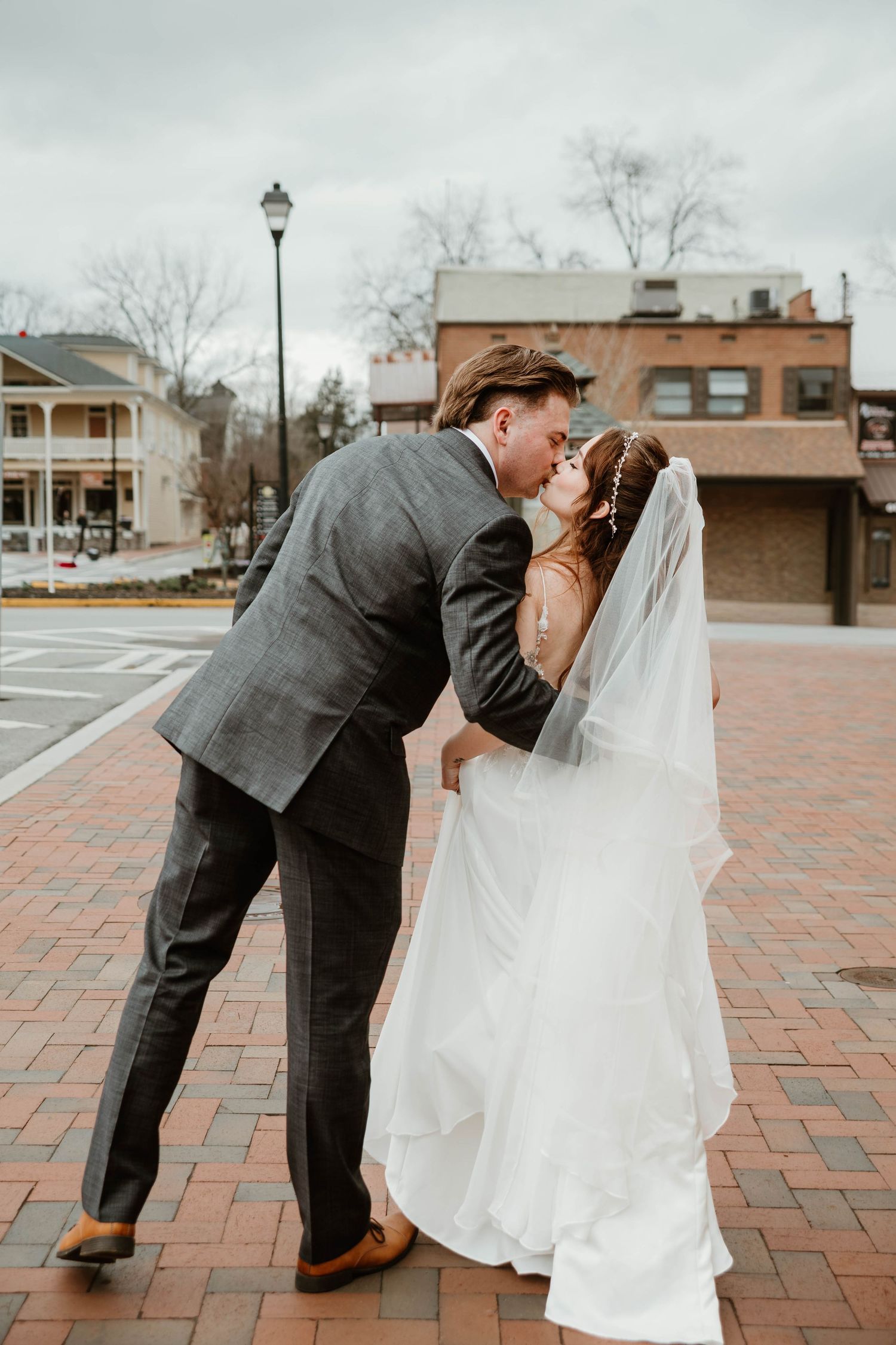 A bride and groom share a romantic kiss on a brick plaza in a small town, she in a white gown and veil, he in a gray suit.