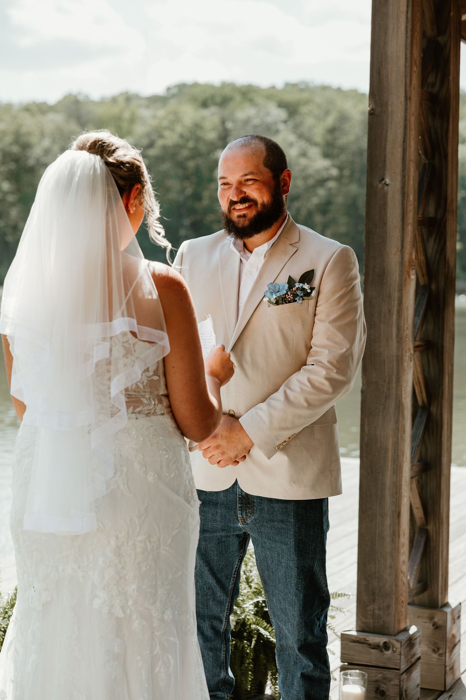 Groom in beige blazer and jeans smiles at bride in lace dress and veil during outdoor lakeside wedding ceremony.
