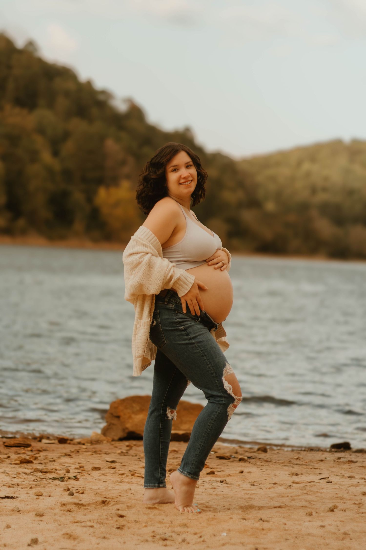 Maternity portrait at sunset by Lake Hartwell in northeast Georgia featuring casual style with cream cardigan and distressed jeans.