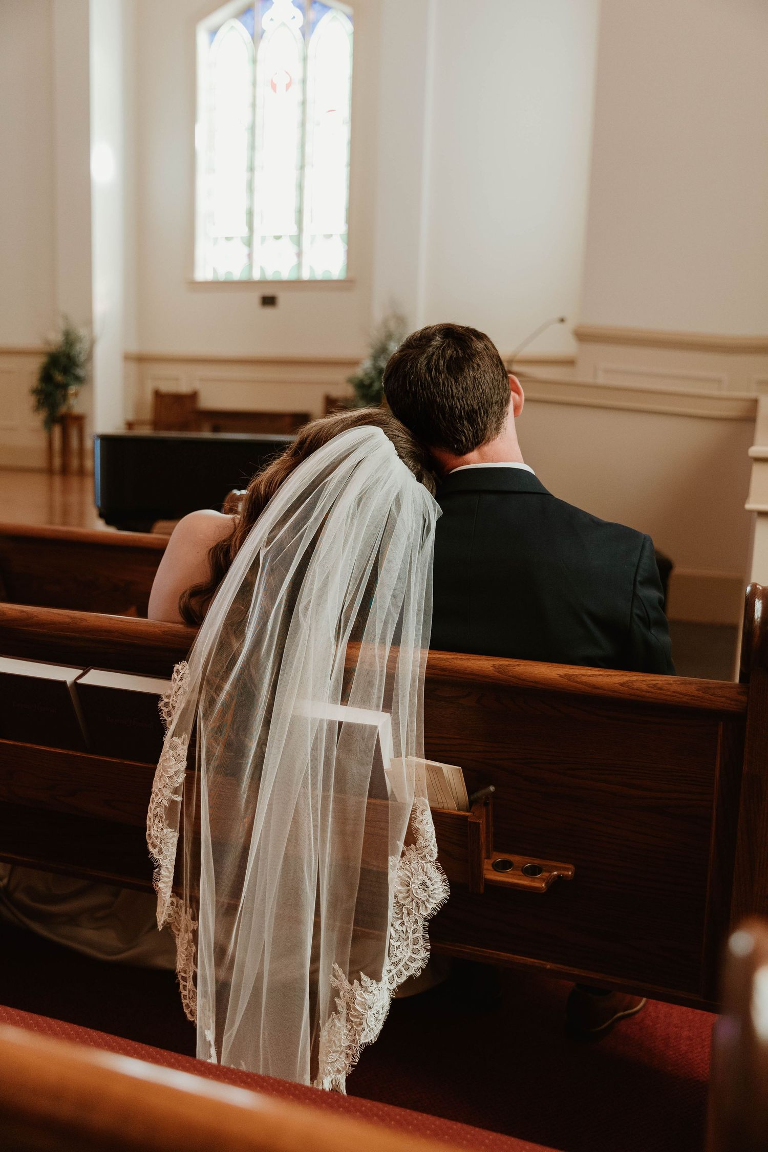 Bride and groom sit together in wooden church pew, bride wearing lace-trimmed veil, stained glass window in background.